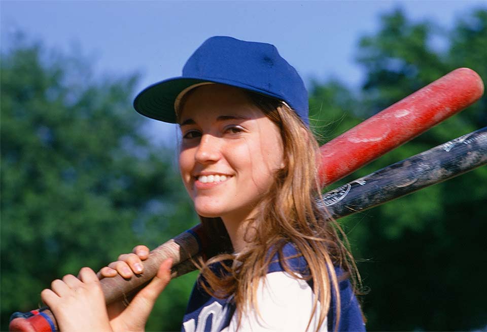 Jan in high school in a softball uniform holding two softball bats over her shoulder