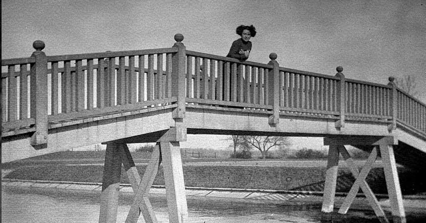 1915 Student leaning over railing on bridge over Lake Marian, sparse trees in background