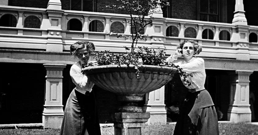 1915 image of two girls posing by flower planter in front of hall