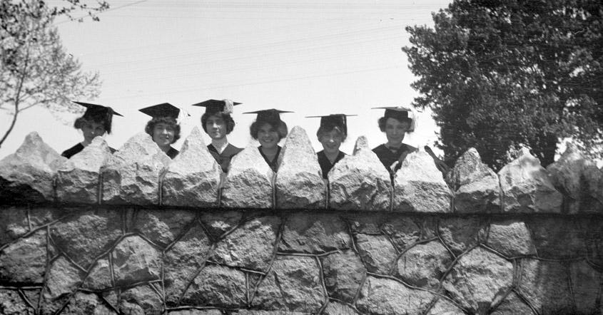 Six graduates in gowns behind stone wall by the Avenue entry